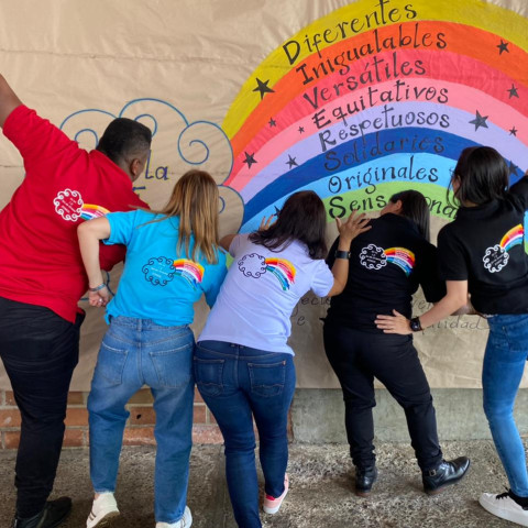 Jóvenes en el pasillo de una escuela pintan un mural con forma de arcoiris de colores que contiene las palabras: diferentes, inigualables, versátiles, equitativos, respetuosos, solidarios, y originales.