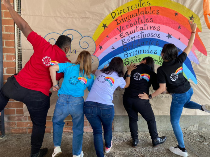 Jóvenes en el pasillo de una escuela pintan un mural con forma de arcoiris de colores que contiene las palabras: diferentes, inigualables, versátiles, equitativos, respetuosos, solidarios, y originales.